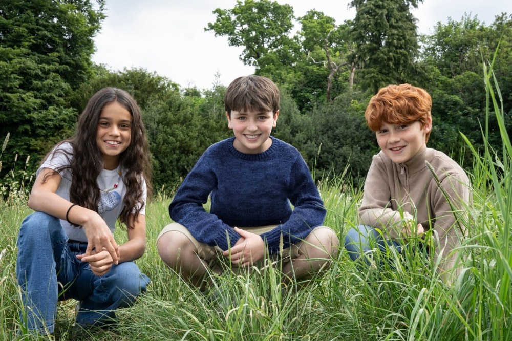 Three young actors sitting together in a grassy area, smiling and posing for the camera, with trees in the background.