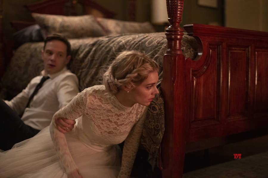 A bride in a wedding dress is crouching under a bed, looking tense, while a man in formal attire sits nearby, appearing anxious.