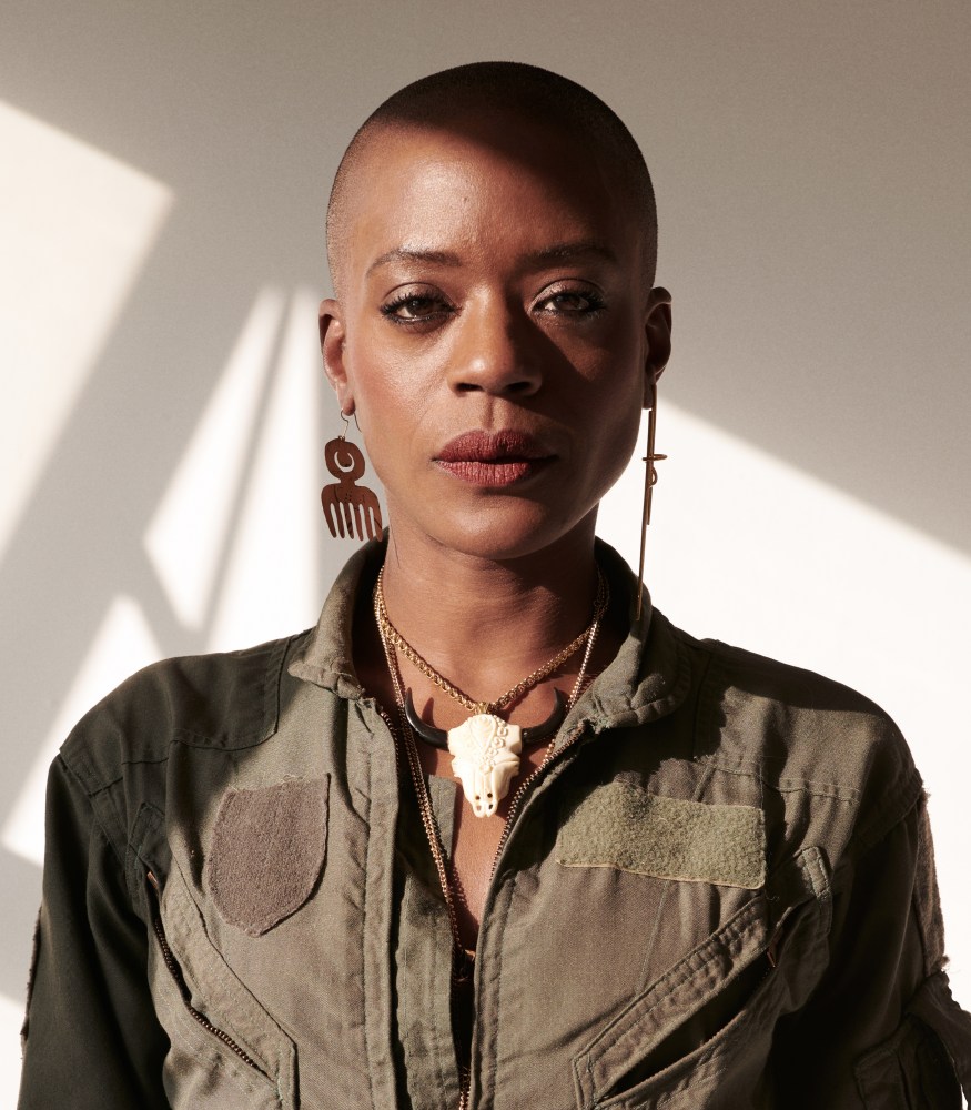 A close-up portrait of a woman with a shaved head, wearing unique earrings and layered necklaces, dressed in a military-style jacket.