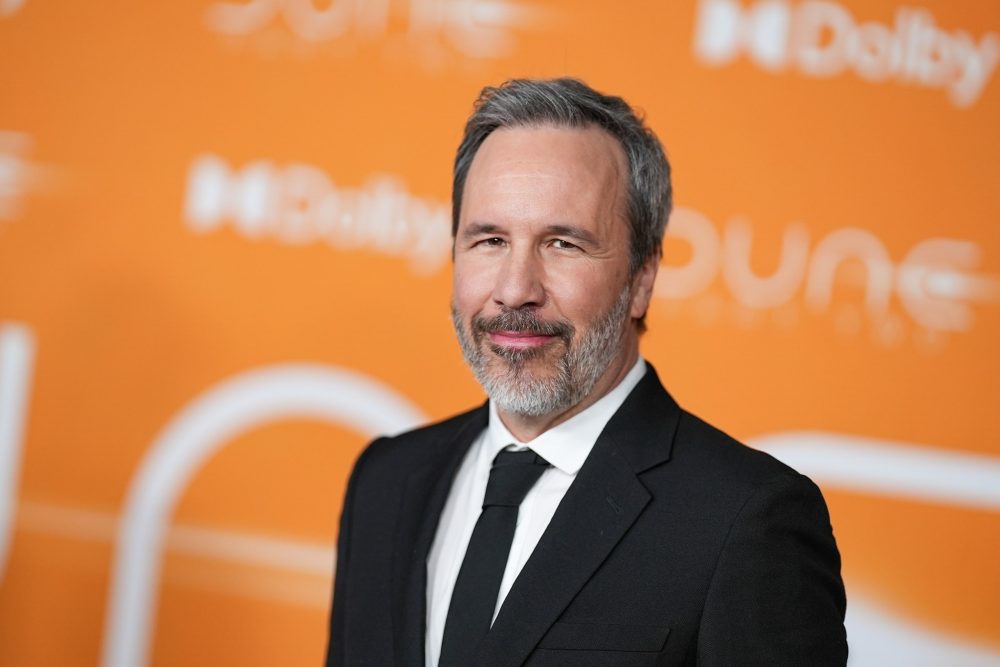 A close-up portrait of a man with a beard and gray hair, dressed in a black suit and tie, smiling against an orange background with the text 'Dune'.