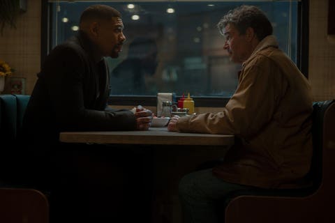 Two men sitting at a diner table, engaged in a serious conversation, with a window view in the background.