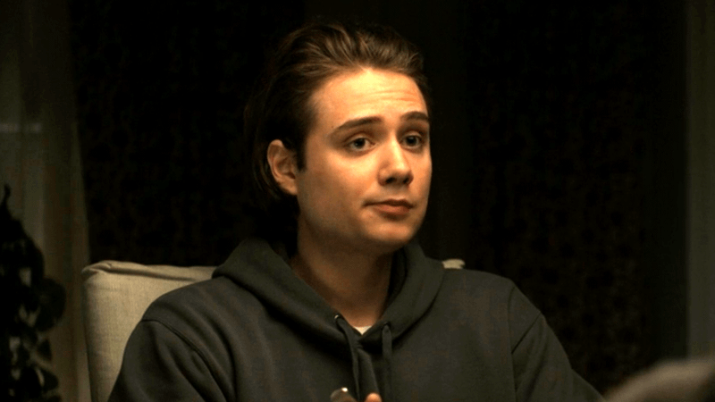 A young man with medium-length brown hair wearing a dark hoodie, sitting at a table with a neutral expression.