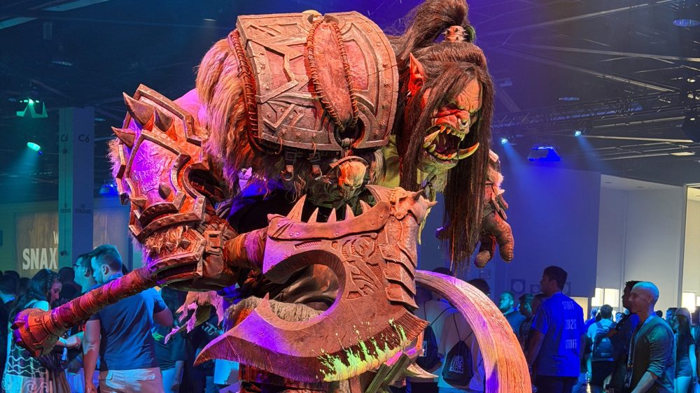A large, detailed orc costume character holding a massive axe, displayed at a convention with a crowd in the background.