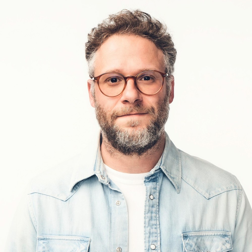 A man with curly hair and glasses, wearing a light blue denim jacket over a white shirt, smiling at the camera against a white background.