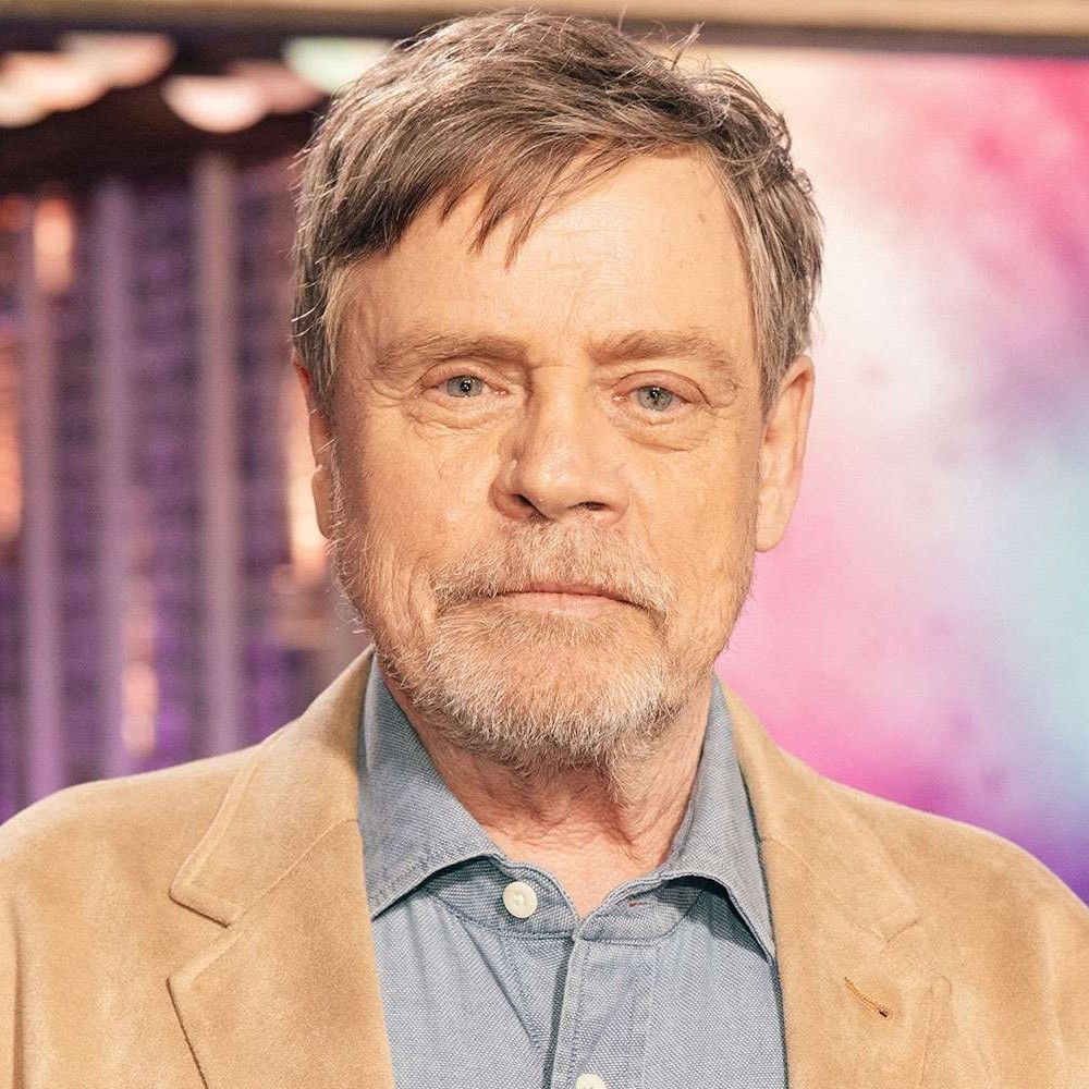 A man with gray hair and a beard, wearing a light tan jacket over a blue shirt, looking directly at the camera with a serious expression.