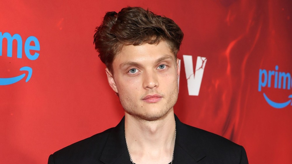 A man with wavy short hair and blue eyes wearing a black jacket, posing against a red background featuring Amazon Prime branding.
