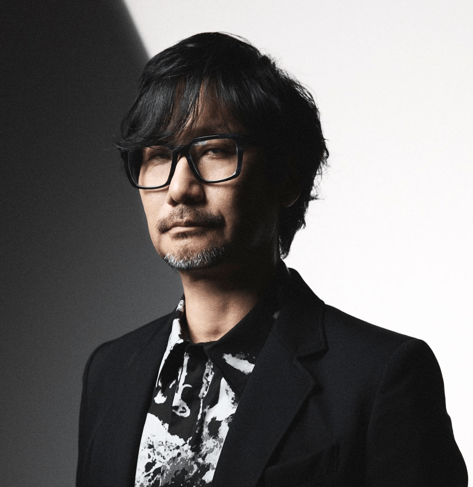 Portrait of a man with glasses and a stylish hairdo, wearing a black blazer over a patterned shirt, set against a light-colored background.