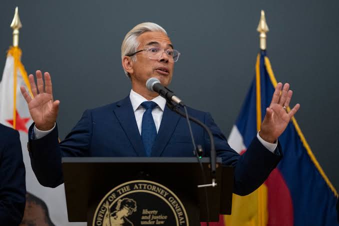 A speaker at a podium with the seal of the Office of the Attorney General, gesturing with both hands while addressing an audience.