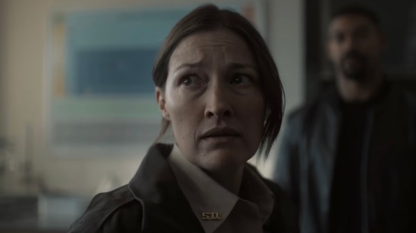 A concerned woman in a uniform looks anxiously over her shoulder, with a man standing behind her in a dimly lit room.