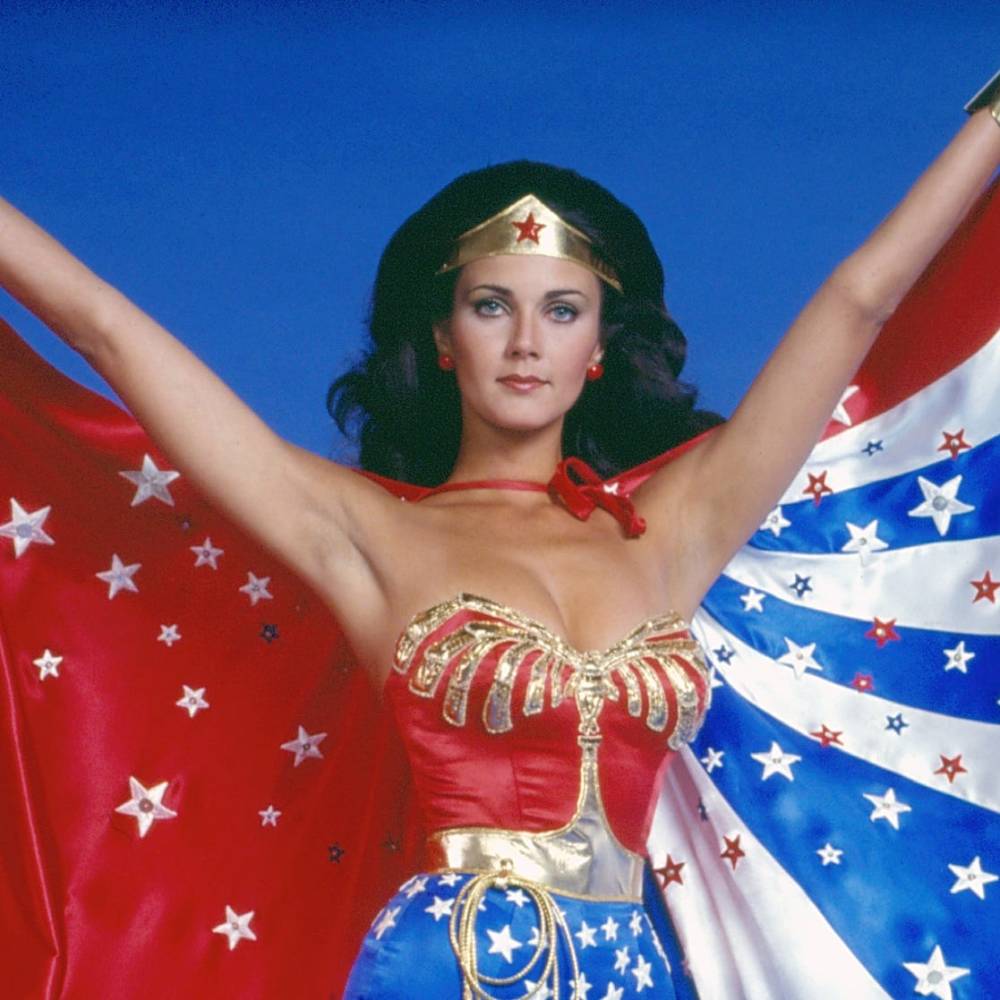 Woman dressed as a superhero with a red and blue costume, gold accents, and a star-patterned cape, posing confidently with arms outstretched against a blue background.