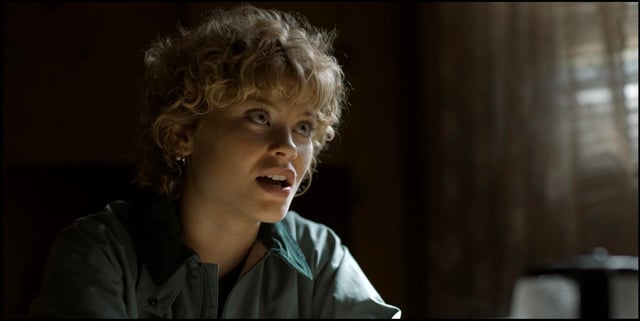 A young person with curly hair wearing a green jacket, sitting at a table and speaking emphatically in a dimly lit room.