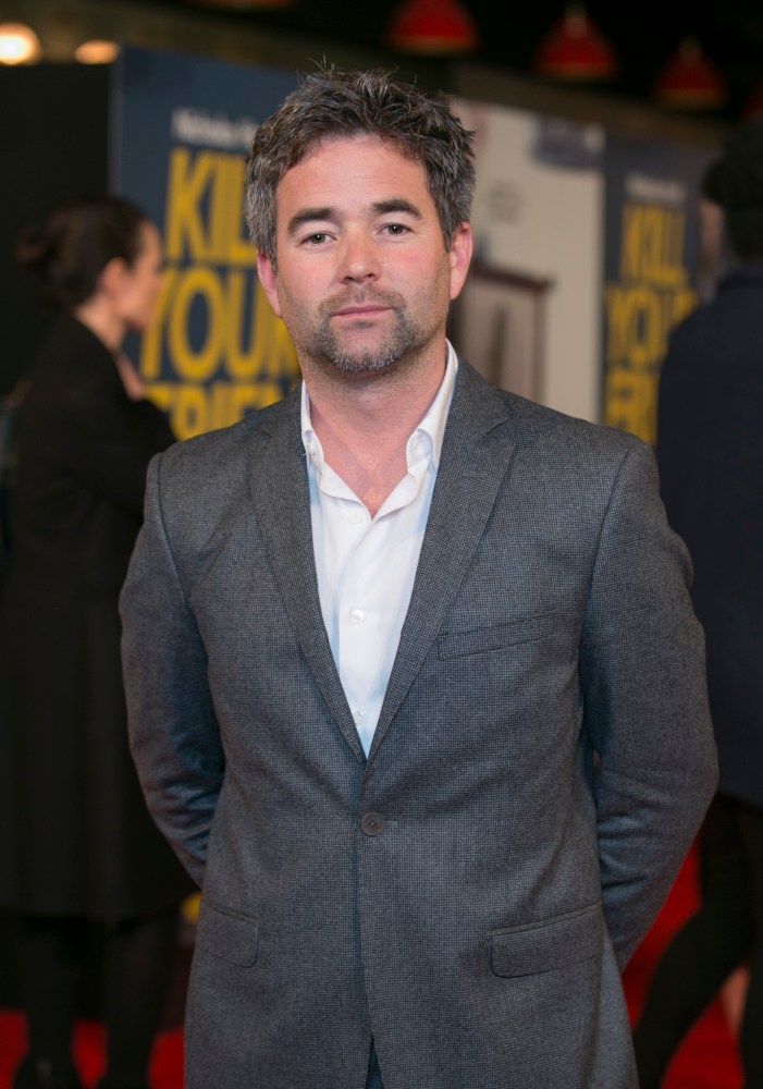 A man wearing a gray blazer and white shirt poses for a photo at a red carpet event, with promotional posters in the background.