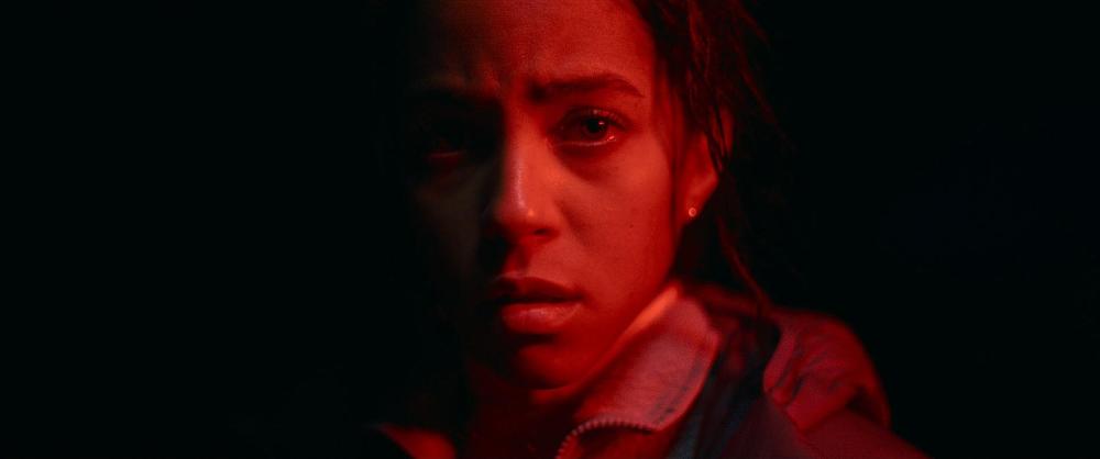Close-up portrait of a woman with a serious expression, illuminated by red light against a dark background.