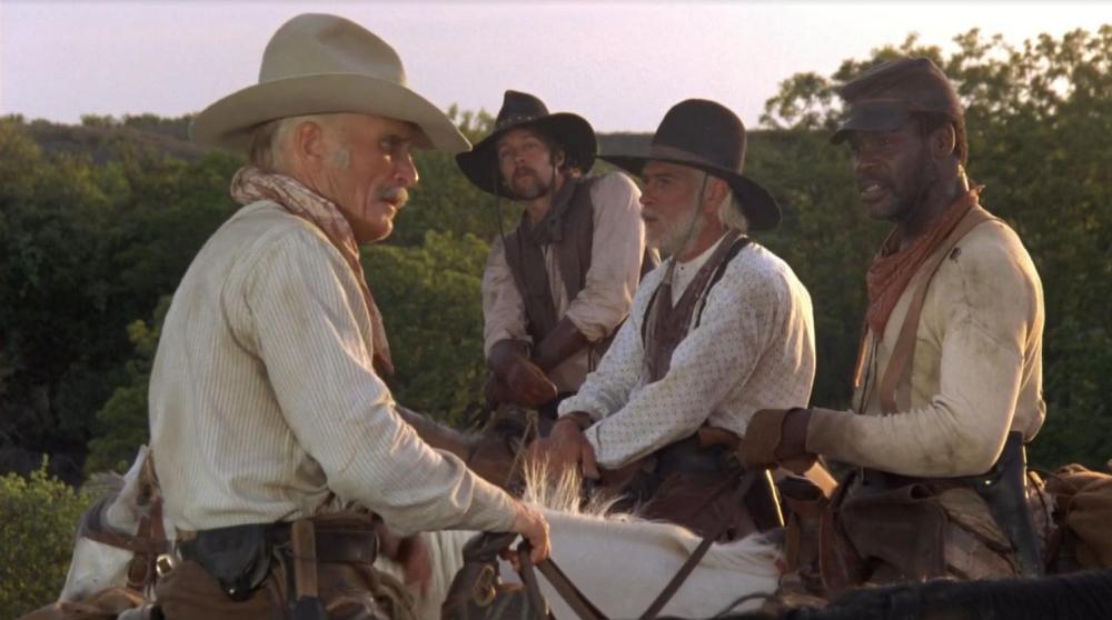 Four cowboys in Western attire engaged in conversation while mounted on horses in a rural outdoor setting.