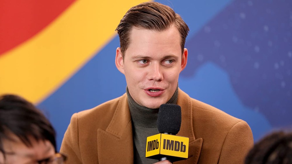 A man wearing a brown coat and green turtleneck speaks into a microphone with an IMDb logo, set against a colorful background.