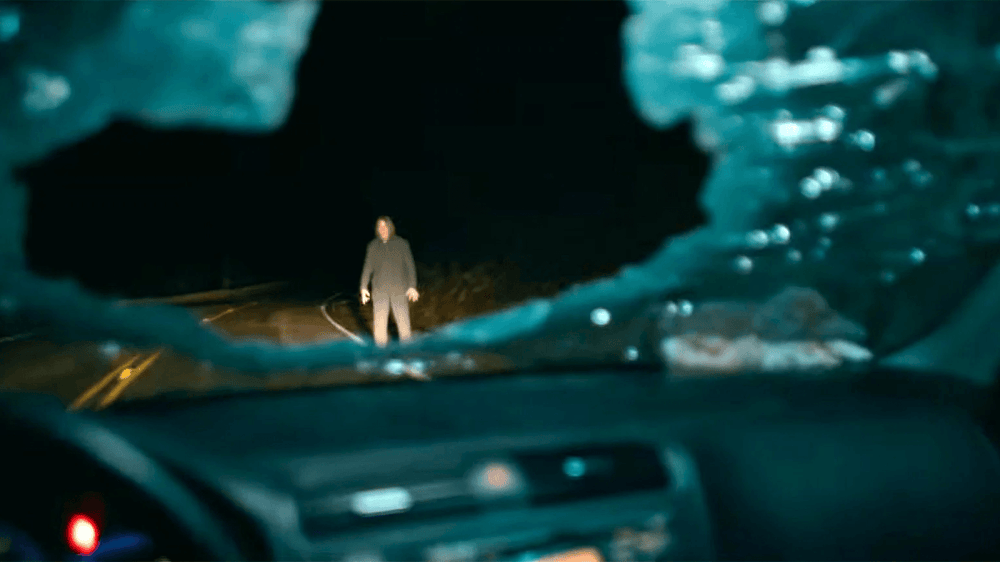 View from a car showing a figure standing in the middle of a dark road, framed by a broken window.