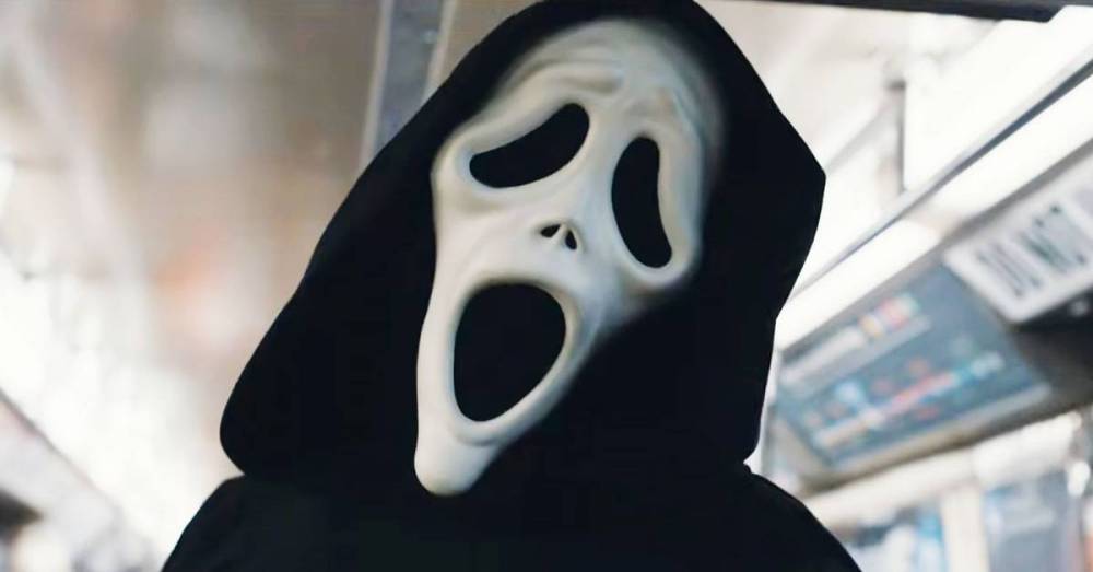 A person wearing a black hooded cloak and a white ghost mask, standing in a subway train.