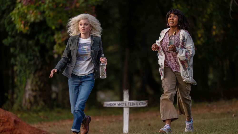 Two women running in a wooded area, one holding a water bottle, with a white wooden cross marker visible in the background.