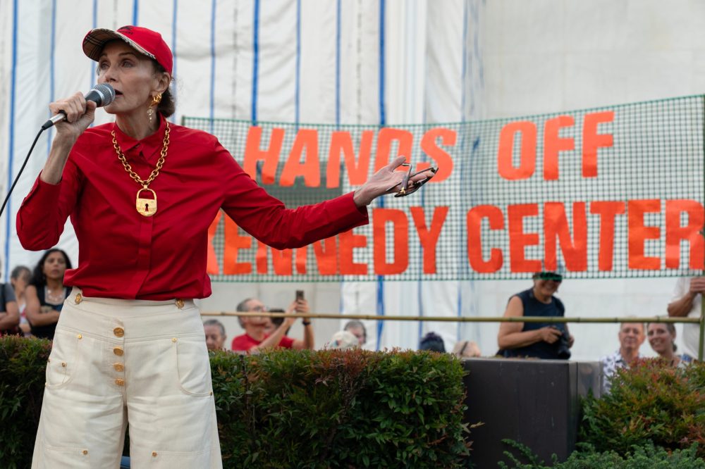 A woman in a red shirt and hat speaks into a microphone at a rally, with a sign reading 'HANDS OFF KENNEDY CENTER' visible in the background.