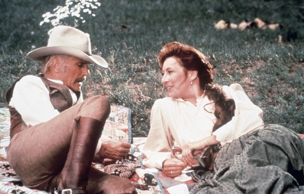 A man wearing a cowboy hat and brown vest sits next to a woman in a white blouse, both relaxed on a picnic blanket in a grassy area.