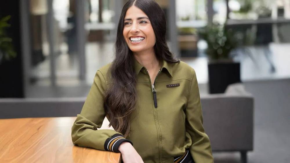 Smiling woman in a green jacket sitting at a wooden table in a modern indoor setting.