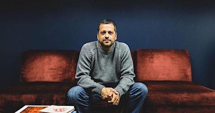 A man sitting on a maroon sofa, wearing a gray sweater and jeans, with a focused expression, against a dark blue wall.