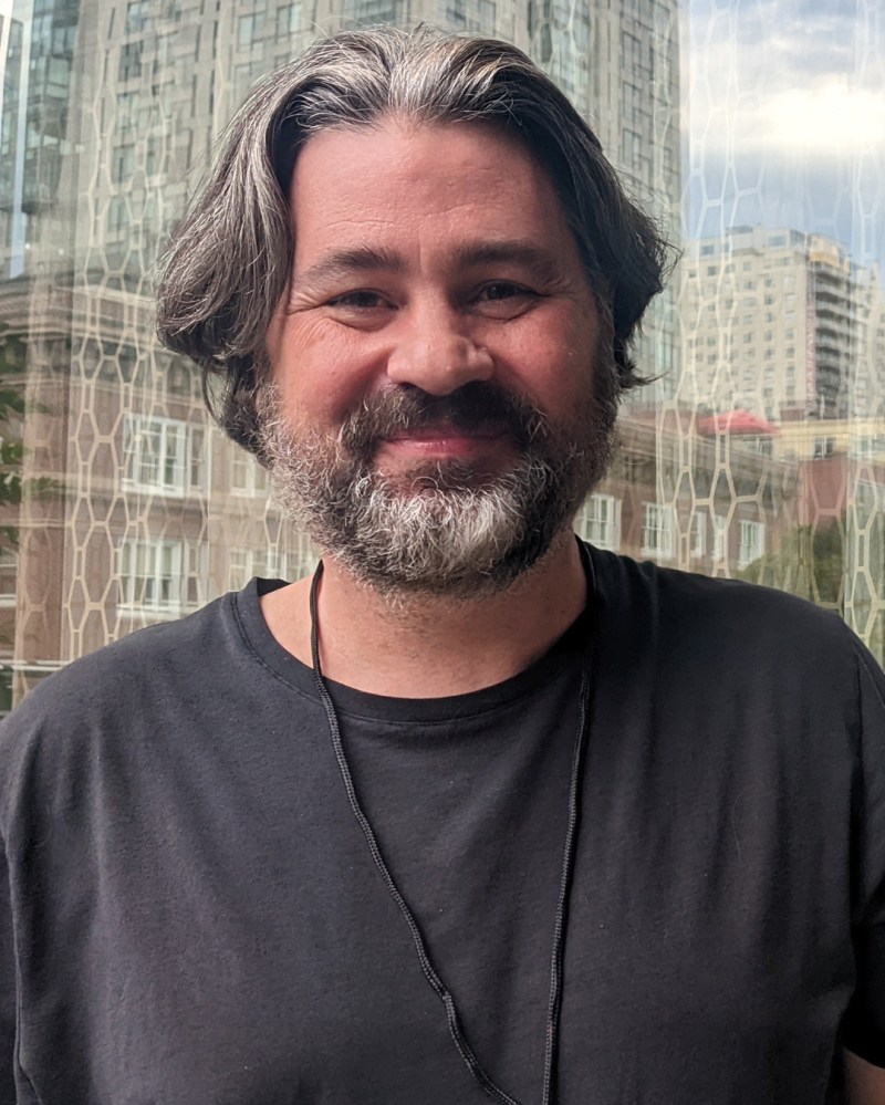 A smiling man with medium-length, wavy gray hair and a beard, wearing a black T-shirt, standing in front of a window with a cityscape in the background.