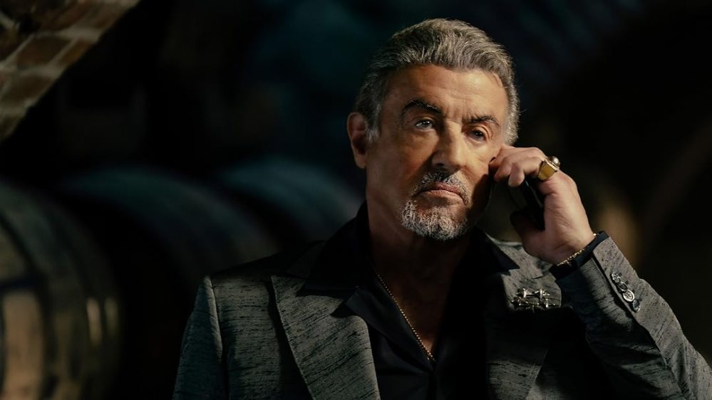 A middle-aged man with grey hair and a beard is wearing a stylish suit and speaking on a phone, appearing serious in a dimly lit environment with barrels in the background.