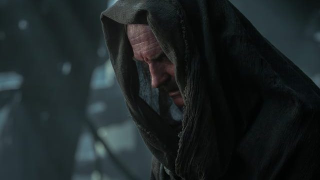 A close-up of a somber, older man wearing a dark hooded cloak, looking downward with a thoughtful expression in a dimly lit environment.