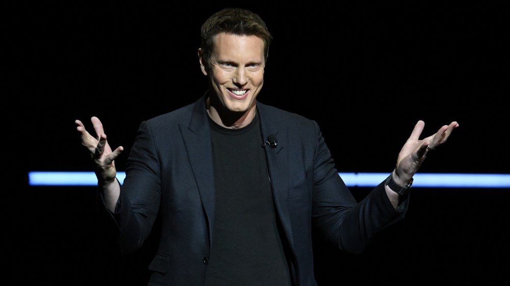 A man in a suit smiles and gestures with his hands while speaking on stage against a dark background.