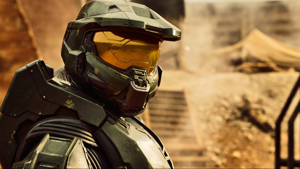 A futuristic armored soldier wearing a helmet with a visor, set against a dusty landscape with steps and a tent in the background.