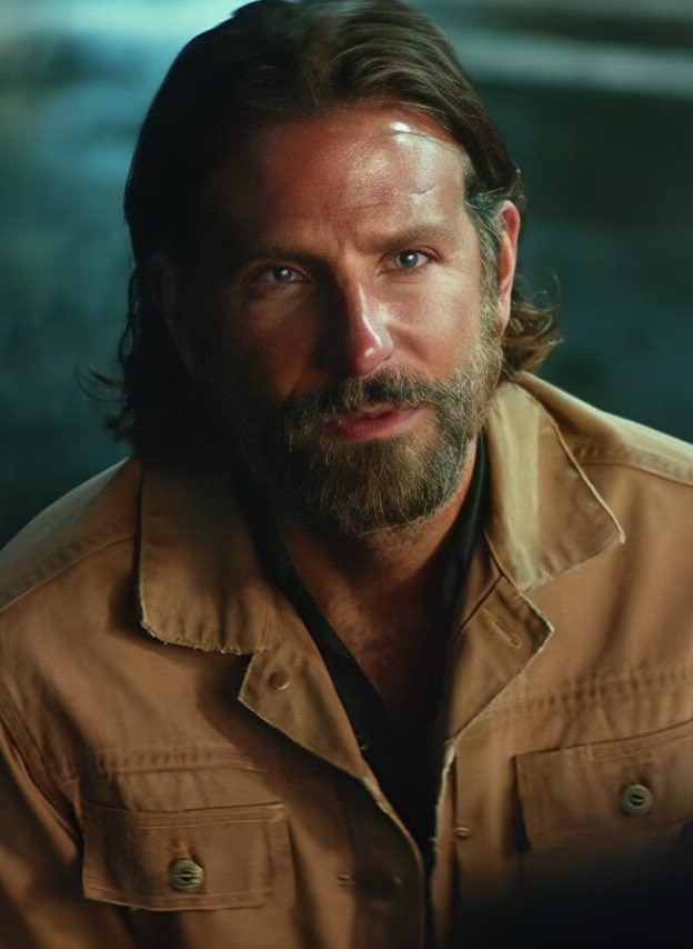 Close-up portrait of a man with long hair and a beard, wearing a tan jacket, looking directly at the camera.