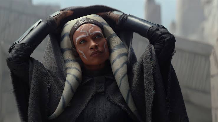 A character with facial markings and a unique headdress is adjusting their hood in a dramatic pose, set against a backdrop of futuristic architecture.
