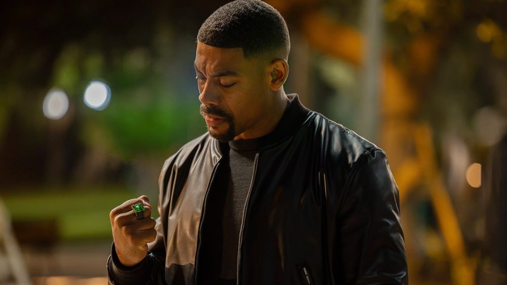 A man in a black leather jacket gazes thoughtfully at a green ring in his hand, set against a blurred outdoor background at night.