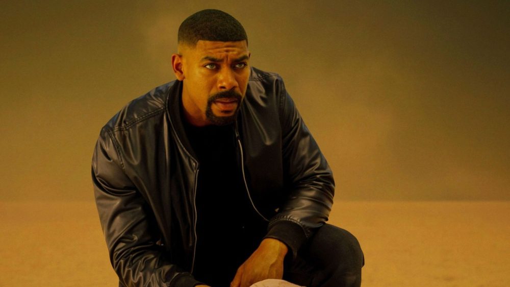 A man in a dark bomber jacket crouching on the ground, with an intense expression, set against a blurred background.