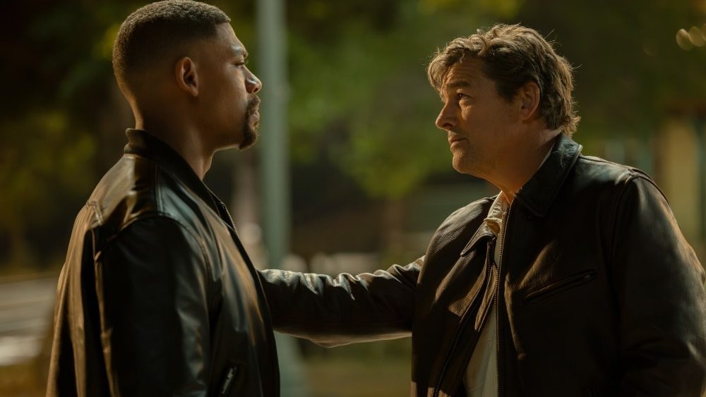 Two men in leather jackets sharing a serious conversation at night, with a blurred background of trees and streetlights.