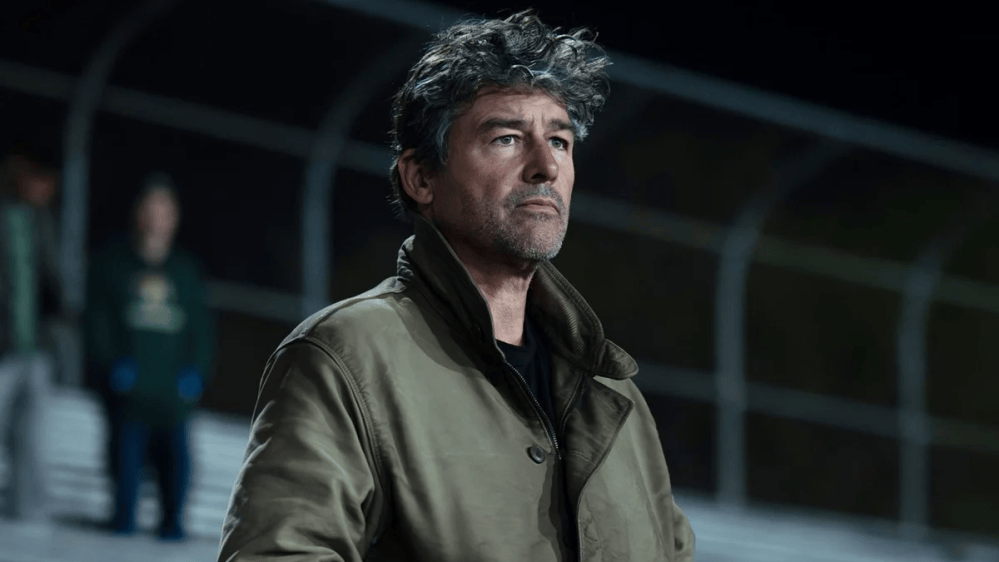 A man with wavy hair wearing a green coat stands pensively in a sports stadium, observing the event.