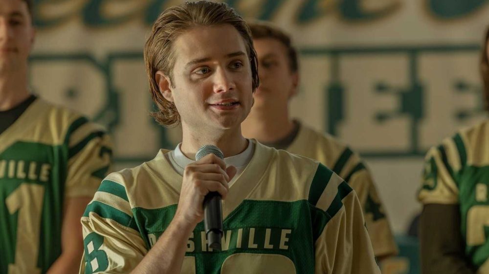 A young man in a football jersey speaking into a microphone, with teammates in jerseys in the background.