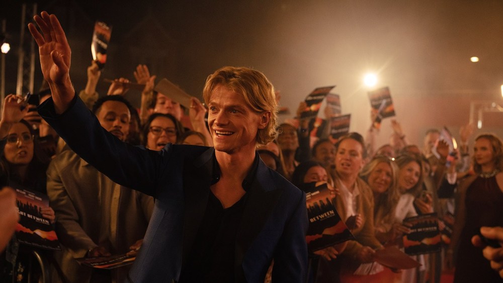 A smiling man in a blue jacket waves to a crowd at a festive event, surrounded by fans holding promotional materials.