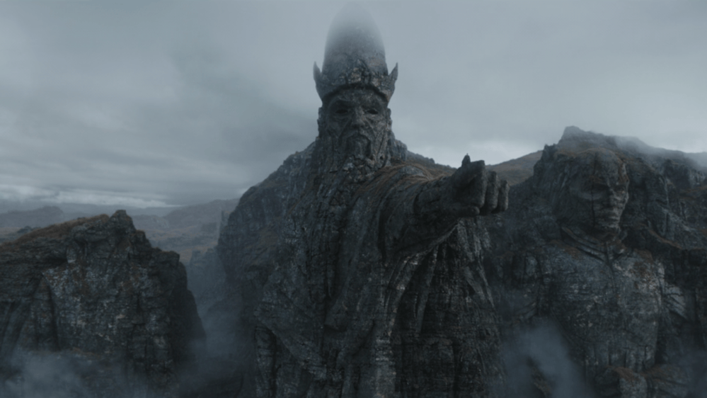 A giant stone statue of a figure with a crown, extending an arm forward, surrounded by misty mountains and rocky terrain.