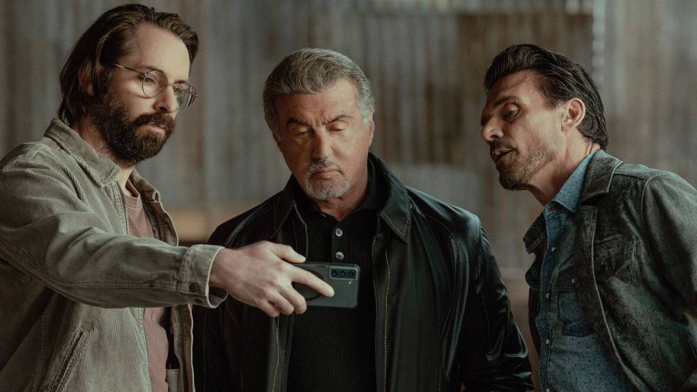 Three men in casual attire are looking at a smartphone together, with one man holding the phone and the others focused on the screen.